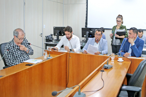 Integrantes da Comissão estão sentados à Mesa apreciando a pauta da reunião. Atrás, há uma servidora de pé, assessorando a reunião