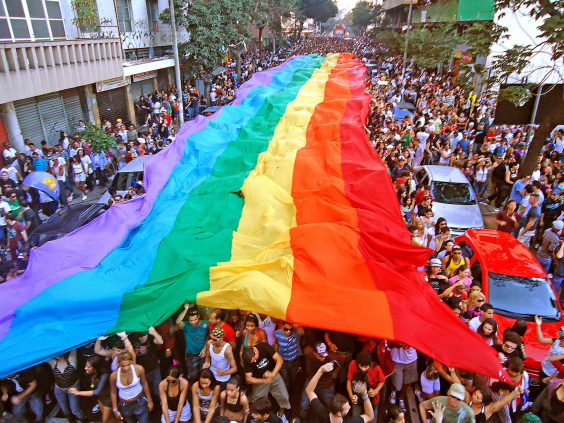 manifestação popular em via pública; pessoas erguem bandeirão com o arco-íris