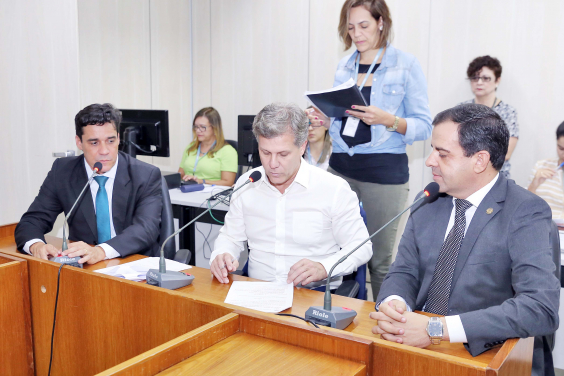 Vereadores estão sentados à Mesa do Plenário apreciando a pauta da reunião; atrás, de pé, uma servidora assessora a reunião