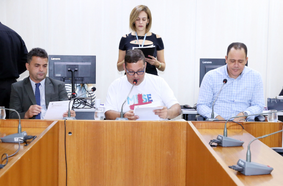 Três integrantes da comissão estão sentados à Mesa, com o presidente no centro; atrás dele, uma servidora está de pé, assessorando os vereadores