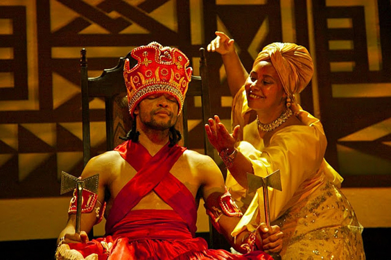 Homem, sentado e trajado como rei, e mulher fazendo uma reverência  em performance teatral