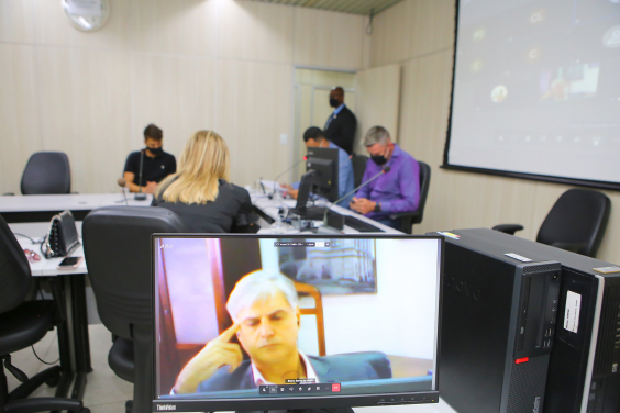 Em primeiro plano, monitor de computador exibe depoente em videoconferência. Ao fundo, parlamentares compõem mesa de reunião presencialmente