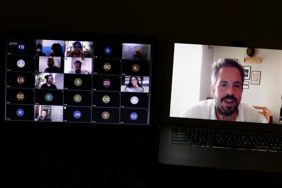 Duas telas de computador: em uma delas, cinco pessoas realizam reunião remota, ne outra, homem participante da reunião está em destaque.