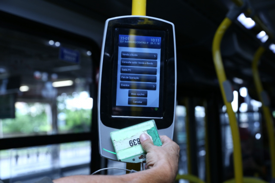 Mão humana passa cartão em máquina de bilhetagem eletrônica, dentro de ônibus, à noite.