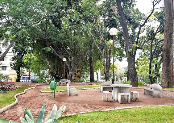 Parque Rosinha Cadar, com duas mess e cadeiras de cimento, cinco bancos, local calçado para transitar e muitas árvores, durante o dia. 