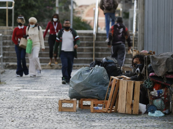 Morador de rua cercado de pertences, sentado em rua de cidade encostado em muro, com  seis cidadãos passando ao fundo, próximo a uma escada de seis degraus, durante o dia.