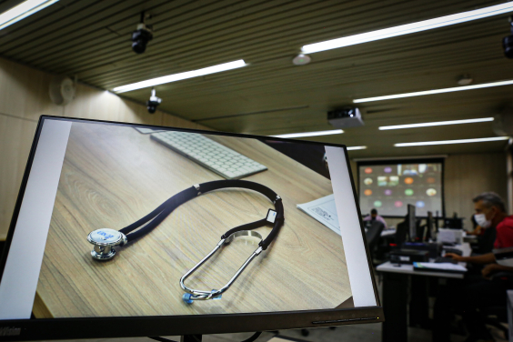 Estetoscópio é exibido na tela do computado em primeiro plano. Ao fundo, vereadores participam de reunião remota