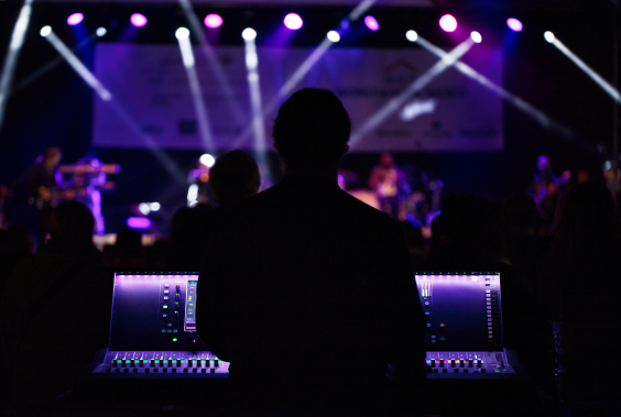Em boate com luz azulada, DJ, de costas, atrás de uma mesa de som, observa pessoas dançando no fundo. 