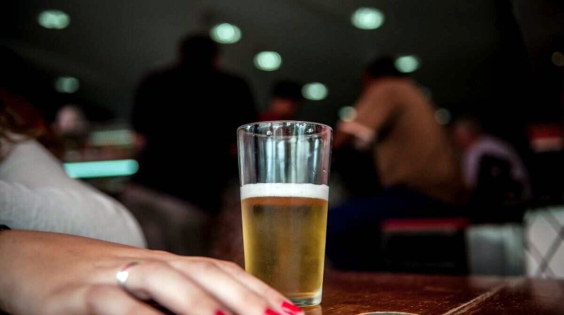 Um copo de cerveja em cima da mesa. Ao lado uma mão de mulher com unhas pintadas de vermelho . A iluminação do teto indica que é um bar ou uma  boate 