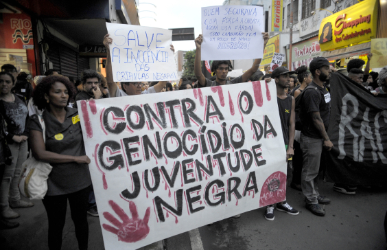 Em rua movimentada, mais de vinte pessoas em passeata seguram cartazes, entre eles dois com os dizeres "Contra o genocídio da juventide negra" e "Salve a inocência das crianças negras".