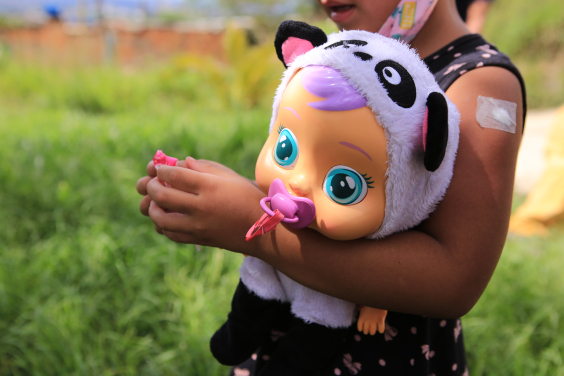 Criança ao ar livre segura uma boneca nas mãos. A boneca, de grandes olhos azuis, usa uma chupeta rosa e uma touca de bichinho
