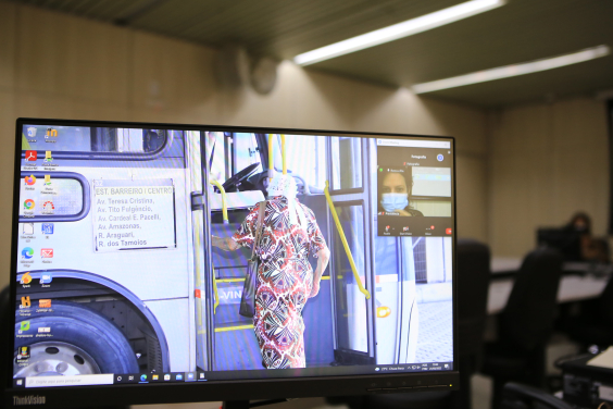 Imagem na tela do computador mostra idosa entrando em um ônibus. Ela tem os cabelos brancos e usa um vestido estampado e uma bolsa a tiracolo