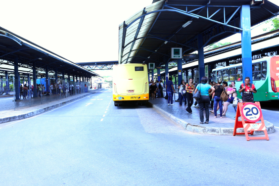 Na Estação Diamante passageiros, em pé, aguardam o ônibus. Estacionado, um ônibus amarelo aguarda a saída