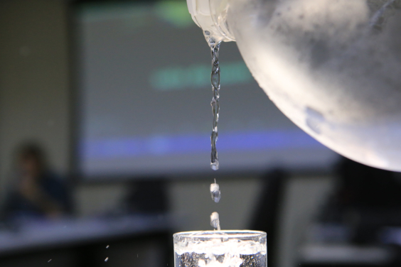Um jarro de água despeja o líquido em um copo transparente . Em segundo plano, a tela do computador com imagem desfocada