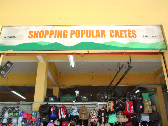 Placa do Shopping Popular Caetés com fundo branco e letreiro em laranja . Abaixo da placa, várias bolsas expostas 