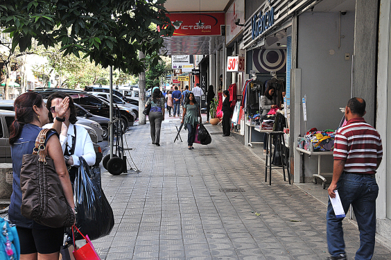 Calçada ocupada por pedestres fazendo compras em lojas diante de um estacionamento repleto de carros. Duas mulheres cheias de sacola conversam paradas na calçada