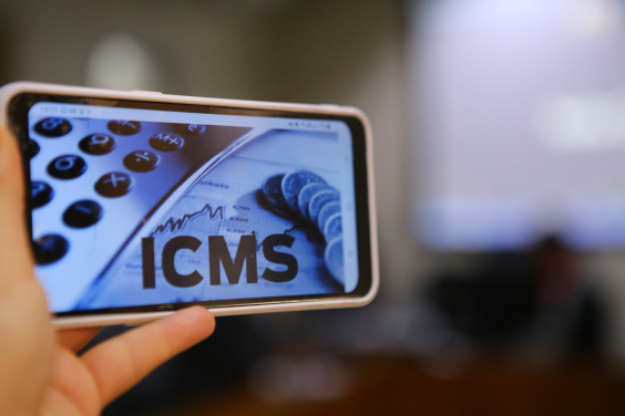 A tela do celular em primeiro plano mostra as teclas de uma calculadora e a  sigla ICMS ao lado de algumas moedas