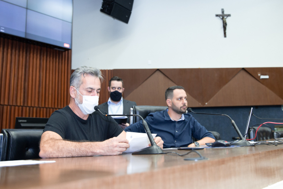 Dois vereadores reunidos, sentados à mesa. Atrás deles, em pé, um assessor da Câmara.