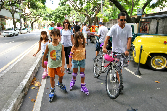 Crianças brincam na rua com patins ao lado de um adulto que está de bicicleta