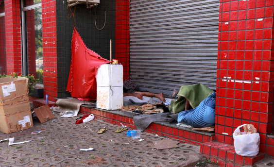 Pessoa em situação de rua , deitado na porta de um estabelecimento comercial fechado. Um pano vermelho está estendido cobrindo uma parte da calçada e há muito lixo na calçada
