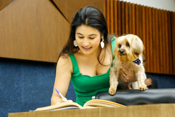 Mulher assina livro enquanto segura cão no braço esquerdo.