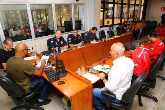 três vereadores reunidos com representantes da Guarda Civil Municipal, do Corpo de Bombeiros Militar de Minas Gerais e dos bombeiros civis, sentados à mesa. Ao fundo, Brigadistas e bombeiros assistem à reunião, sentados.