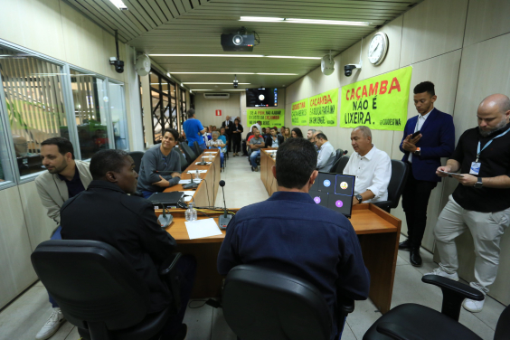 Sala de reuniões com muitos homens e mulheres sentados e de pé, cartazes com dizeres como "Caçamba não é lixo" não paredes.