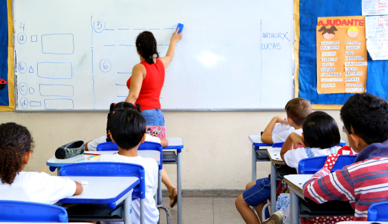 Professora, de costas, escreve em quadro branco; atrás delas, seis crianças, sentadas em carteiras, a observam.