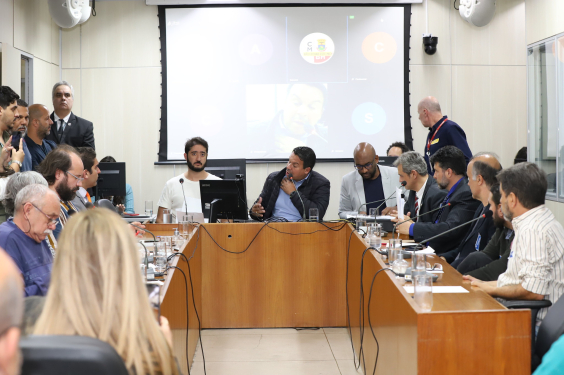 Seis parlamentares e quatro pessoas sentadas à mesa, em uma sala cheia de cidadãos, durante audiência pública.