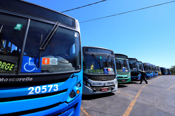 Dez ônibus enfileirados, estacionados em diagonal, com homem verificando a parte da frente de um deles, durante o dia. 