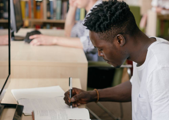 Jovem negro faz anotações em caderno, sentado à frente de computador.