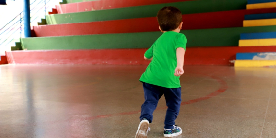 De costas, criança caminha em direção à escada com degraus coloridos, durante o dia.