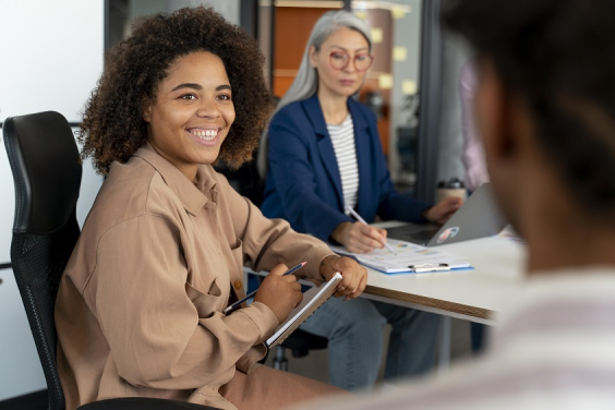Mulher negra sentada em uma cadeira faz anotações em caderno. Atrás dela está uma mulher mais velha de cabelos grisalhos longos, também fazendo anotações. À frente dela, de costas e desfocada, está uma pessoa em pé, ensinando.