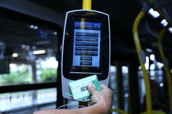 Na foto uma mão encosta um cartão na máquina de cobrança de passagem de um ônibus