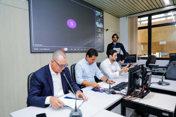 Vereadores estão sentados no plenário. O telão atrás deles mostra a videochamada com parlamentares presentes virtualmente.