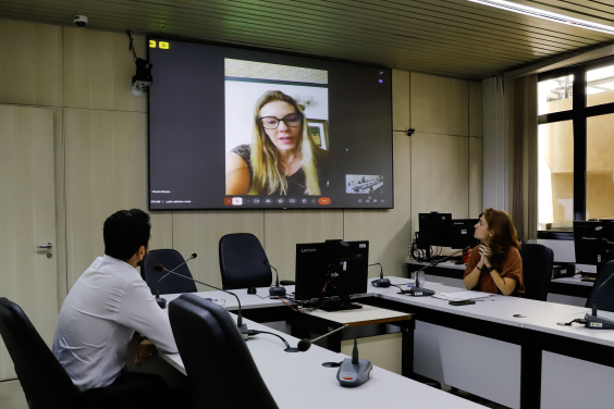 Luiza Dulci e Uner Augusto estão assentados no Plenário Helvécio Arantes. O telão à frente deles mostra Flávia Borja em chamada virtual.