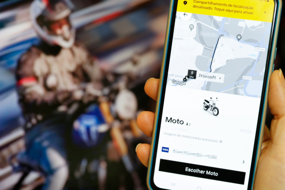 Em primeiro, plano, celular com aplicativo de corrida por moto. Em segundo plano, motociclista