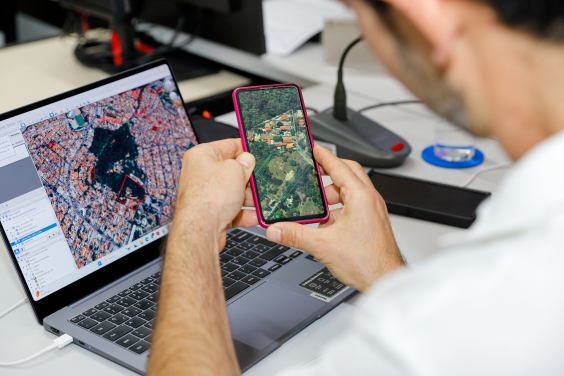 homem com o celular na mãe e o computador à frente, ambos com imagem geográfica de satélite