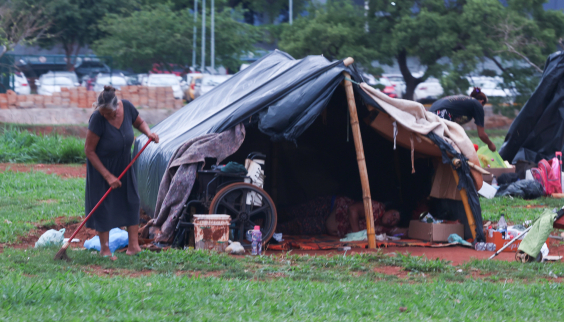 Mulher em situação de rua varre terreno ao lado de cabana improvisada com lona