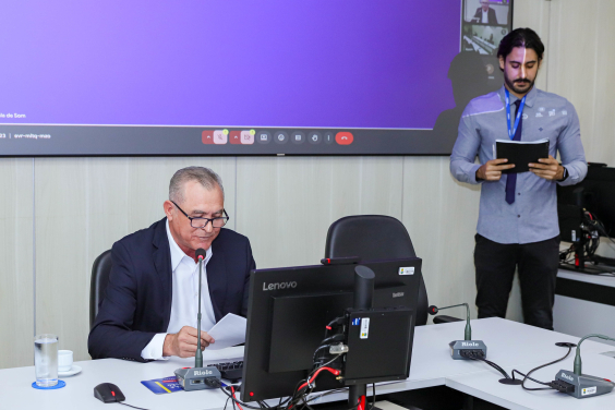 Imagem de vereador segurando a pauta, sentado atrás da mesa de reunião.