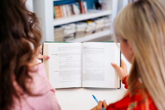 Duas mulheres de costas leem livro didático