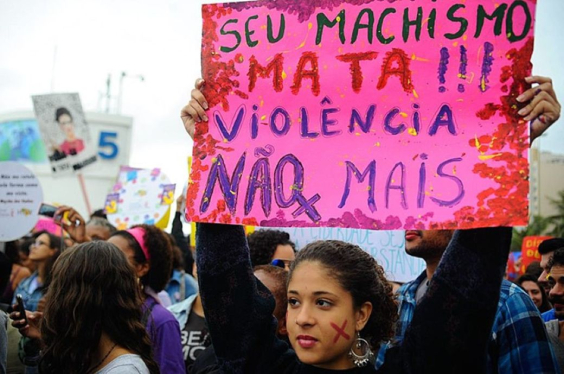 Mulher segura cartaz com os dizeres: "Seu machismo mata!!! Violência não mais"
