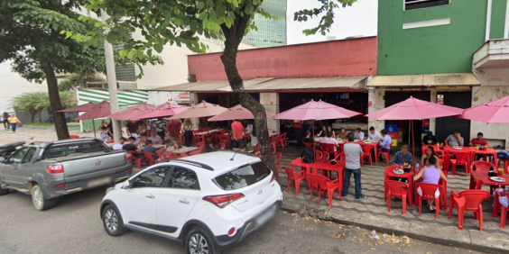 Bar com pessoas sentadas em mesas nas calçadas e carros estacionados na rua, durante o dia.