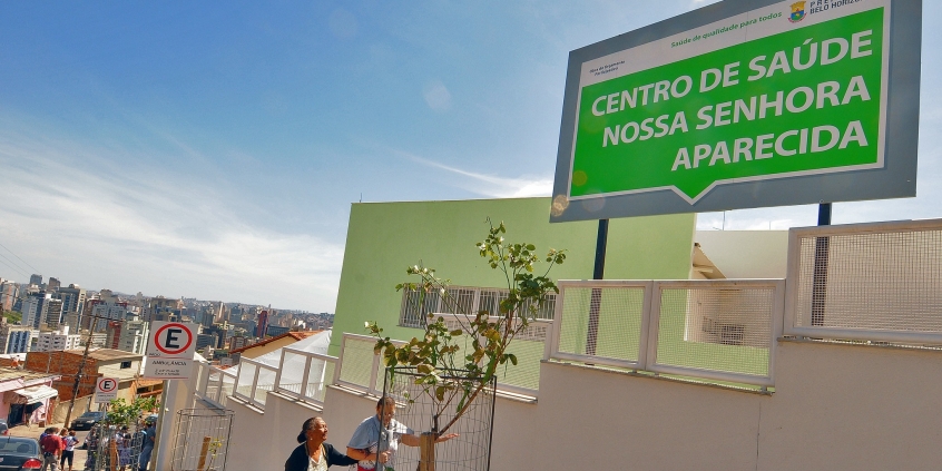 Fachada do Centro de Saúde Nossa Senhora Aparecida com placa indicativa. Rua em declive. Dua mulheres na porta