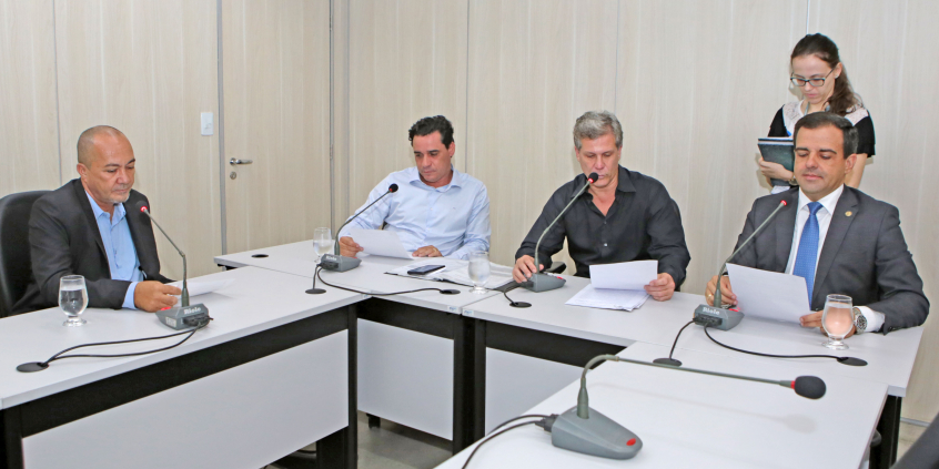 Integrantes da Comissão estão sentados à Mesa apreciando a pauta da reunião; no canto direito da foto aparece uma servidora com uma prancheta na mão