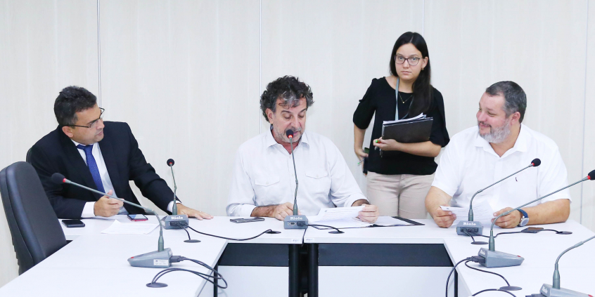 Integrantes da Comissão estão sentados à Mesa apreciando a pauta da reunião. Atrás deles, de pé, uma servidora assessora o presidente.