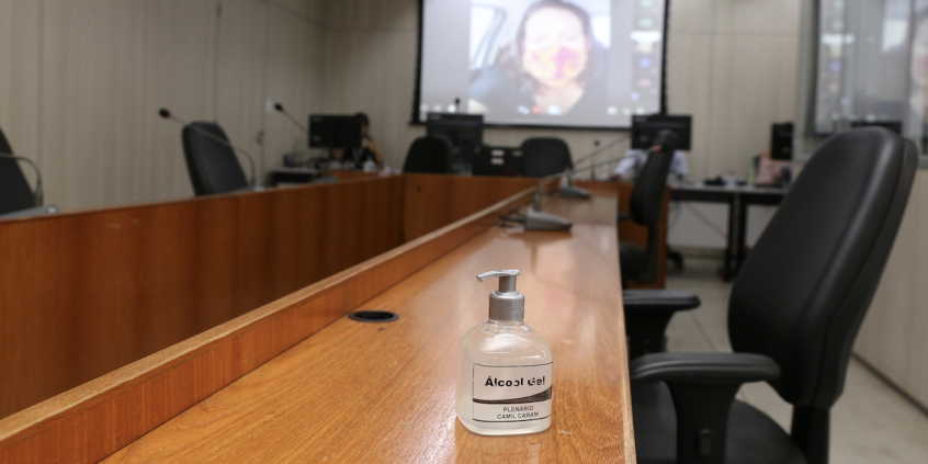 em primeiro plano, vidro de álcool em gel sobre a mesa; cadeiras vazias. Ao fundo, telão exibe reunião em videoconferência