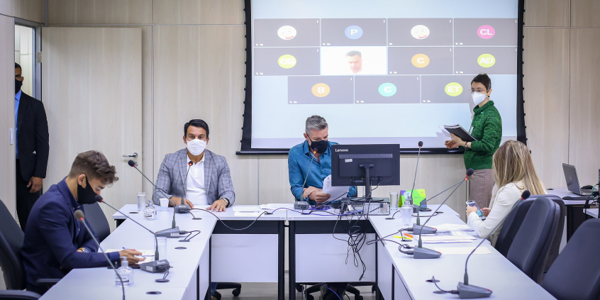 sala de reunião com quatro vereadores sentados distantes um do outro e ao fundo tela de tv com participantes remotos