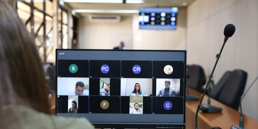 Monitor de computador exibe vereadores em videoconferência. Ao fundo, cadeiras vazias do Plenário Camil Caram