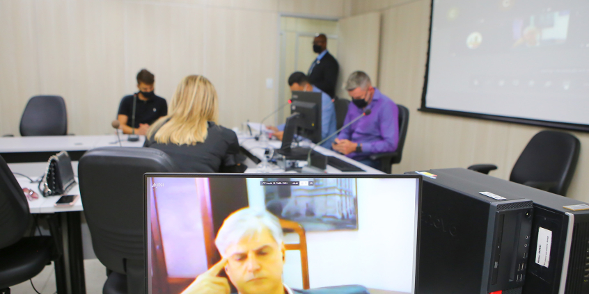Em primeiro plano, monitor de computador exibe depoente em videoconferência. Ao fundo, parlamentares compõem mesa de reunião presencialmente